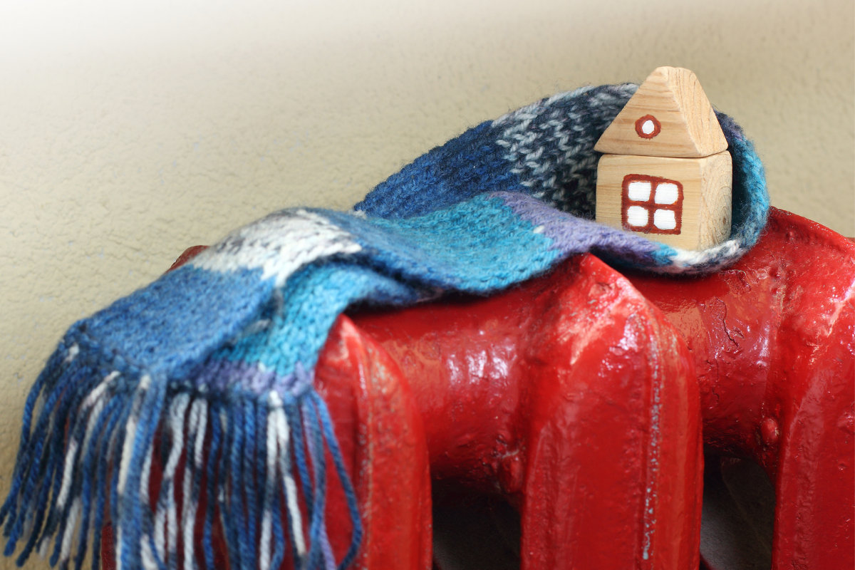 Energy Efficient Heating - Concept feature image from Canva, of a wooden toy block house wrapped in a knitted scarf, perched on top of an old fashioned red metal radiator.